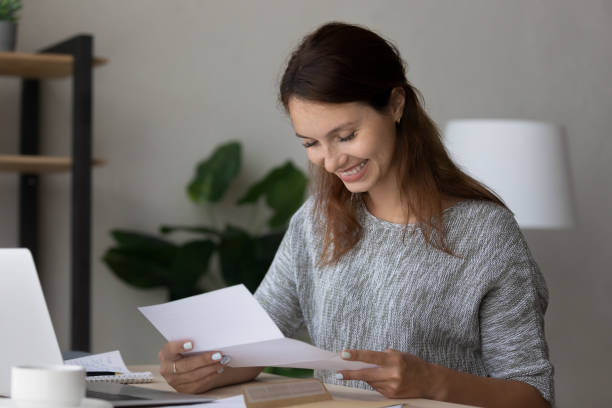 Happy millennial woman reading paper correspondence letter, feeling excited by good news, getting last mortgage payment notification or invitation for event, sitting at home office workplace.